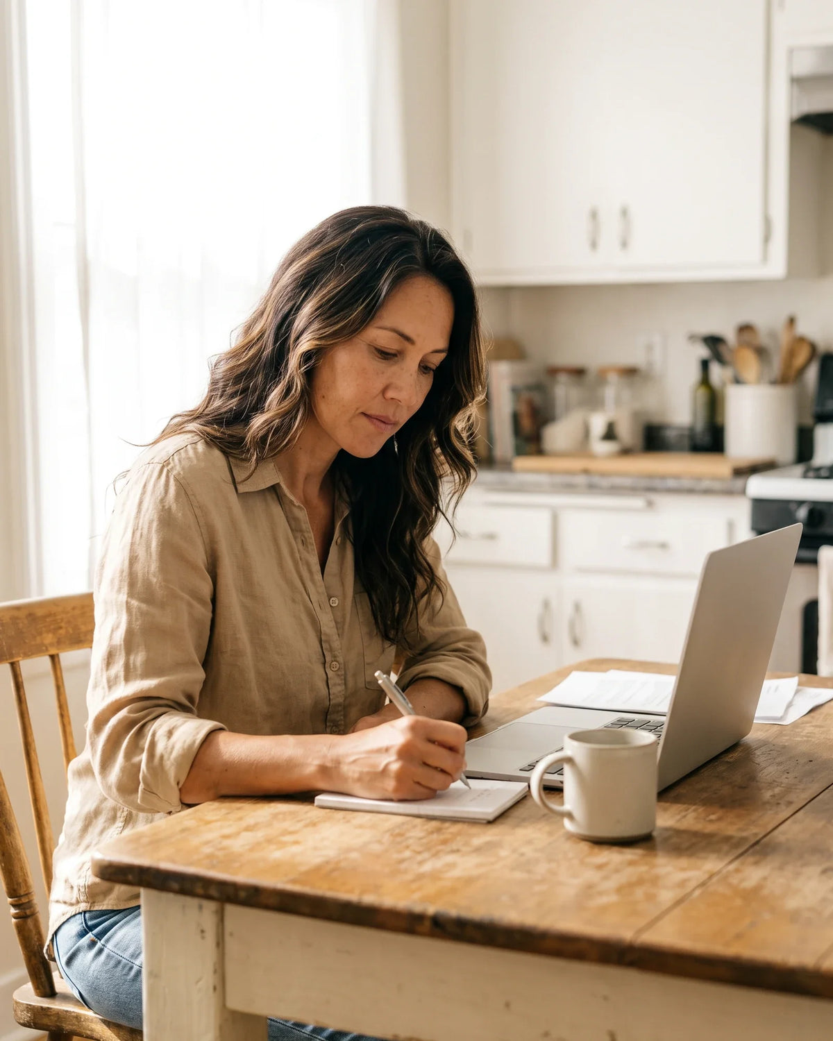 A woman writes in a notebook at a rustic wooden table with a mug and laptop in morning light.