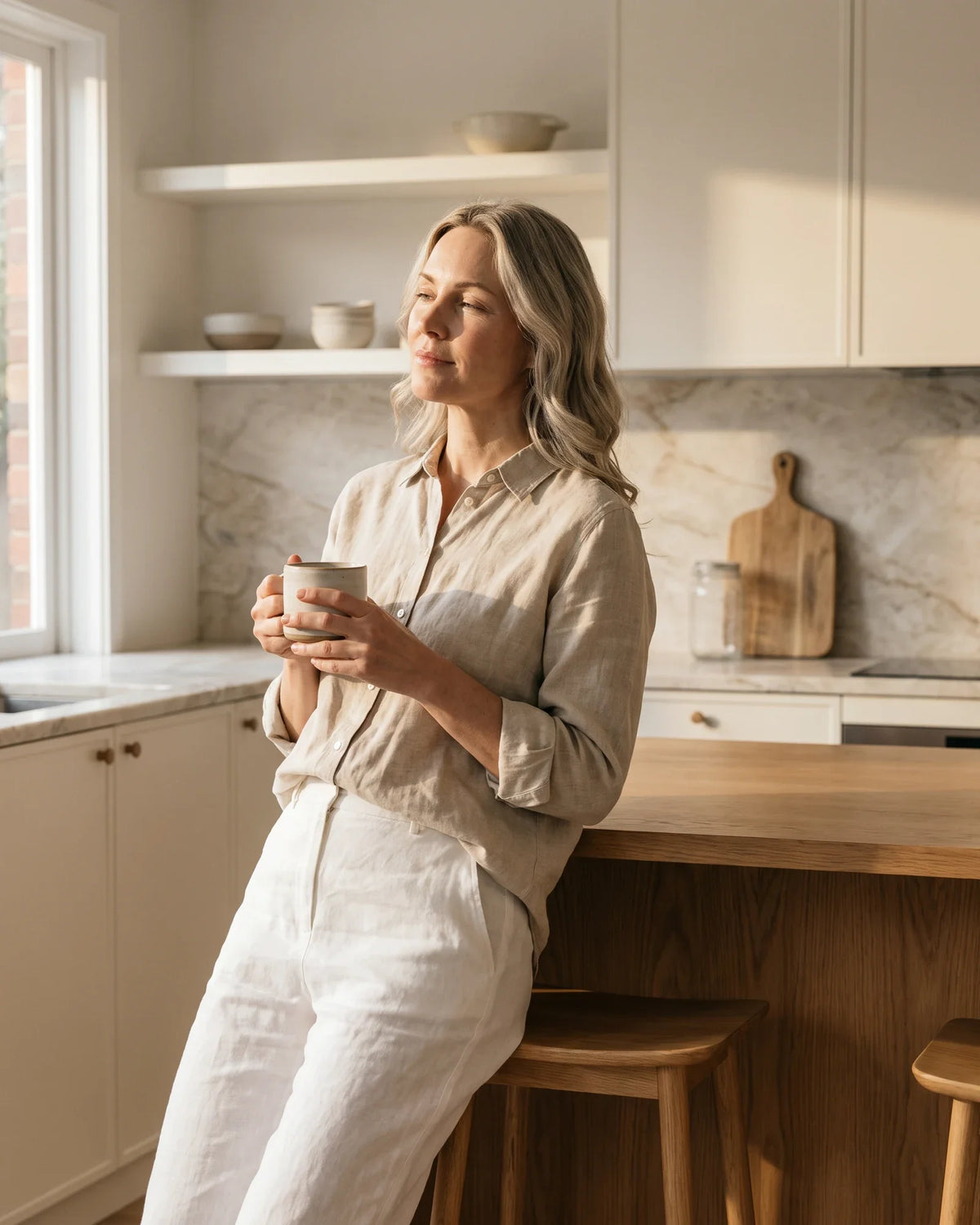 A woman leans against a kitchen counter holding a mug in morning light.