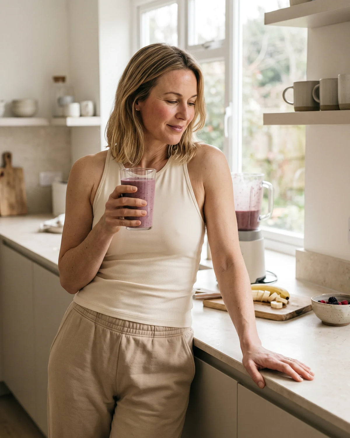A woman leans on a bright kitchen counter holding a smoothie.
