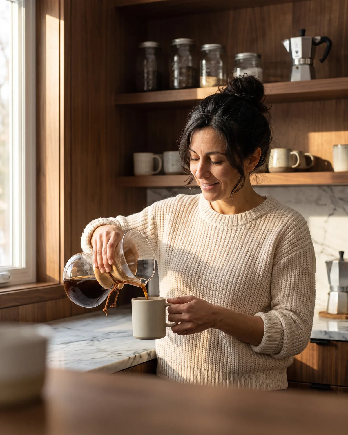 A woman smiles as she pours coffee from a glass carafe into a mug in a warm kitchen.