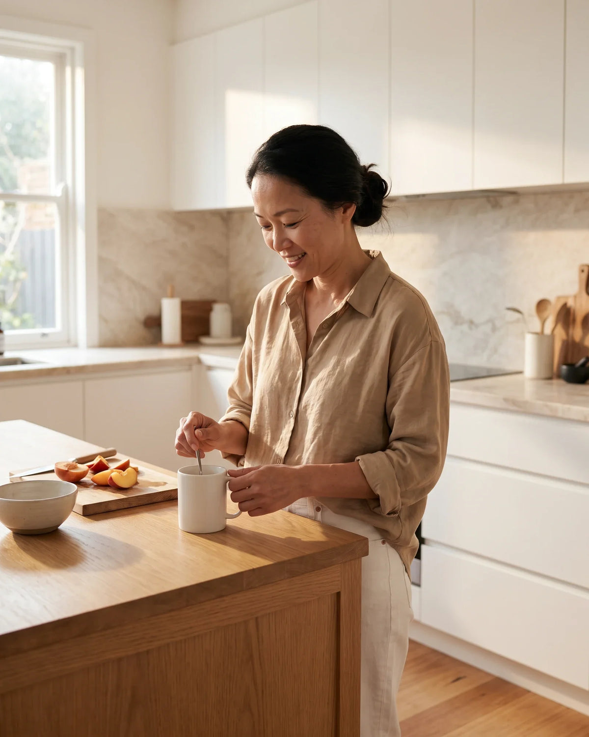 A woman smiles as she stirs a mug at a bright kitchen counter.