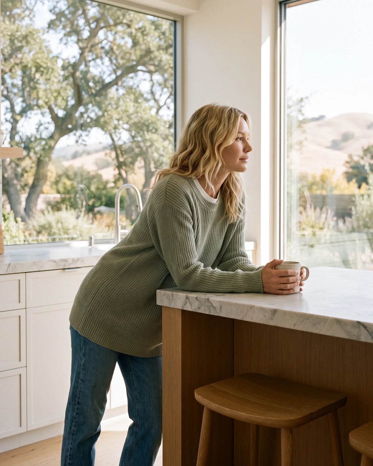 A woman leans on a kitchen counter with a mug, looking out at morning hills.