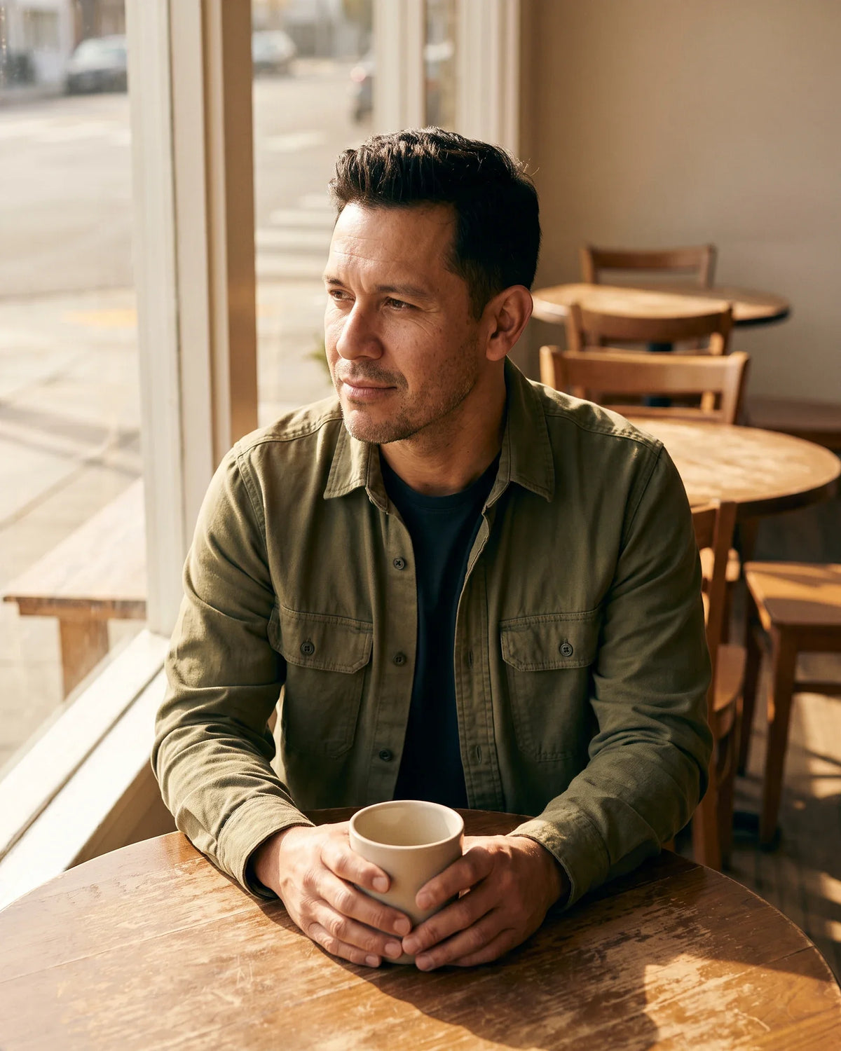 A man holds a mug at a sunlit cafe table and looks out the window.