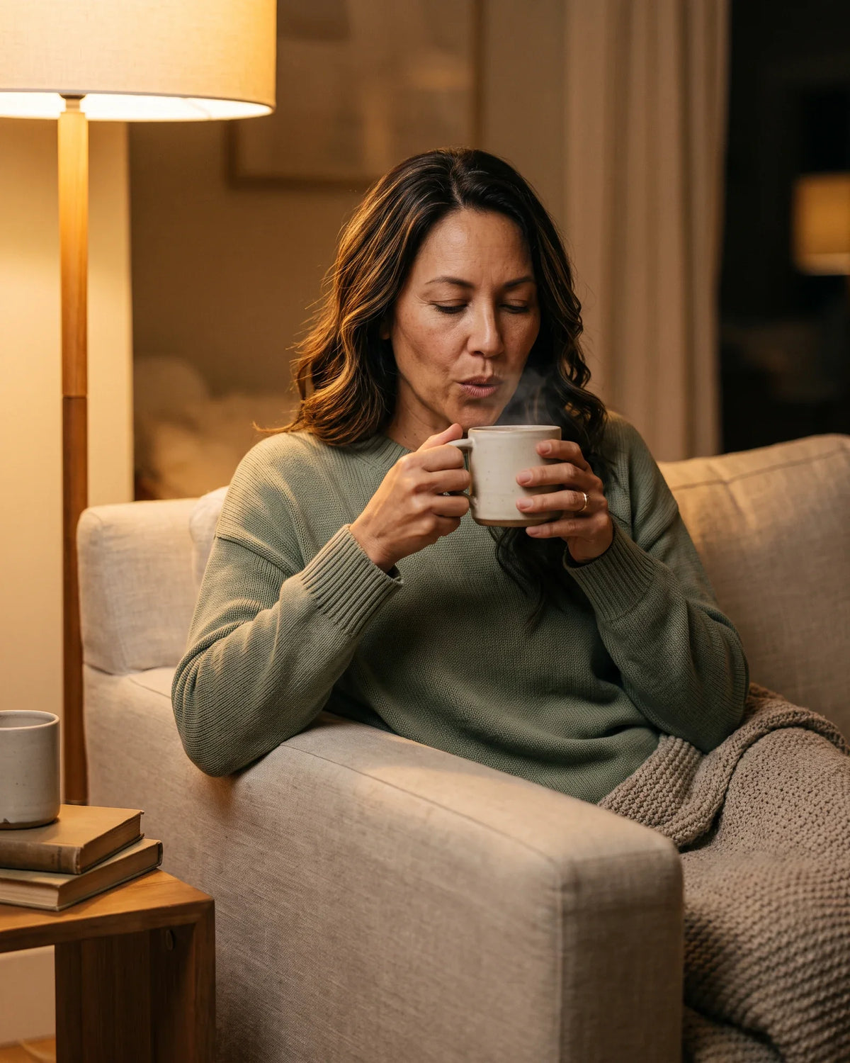 A woman in a green sweater blows on a steaming mug on the sofa.