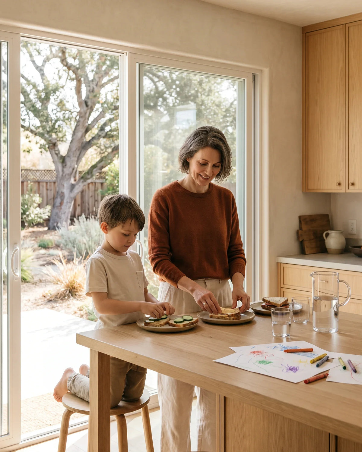 A mother and young son share lunch and drawings at a sunny kitchen counter.