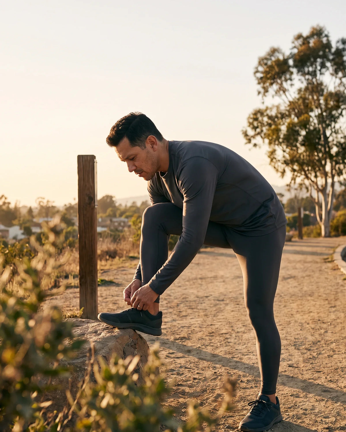 A man ties his running shoe on a rock at the start of a hillside trail.
