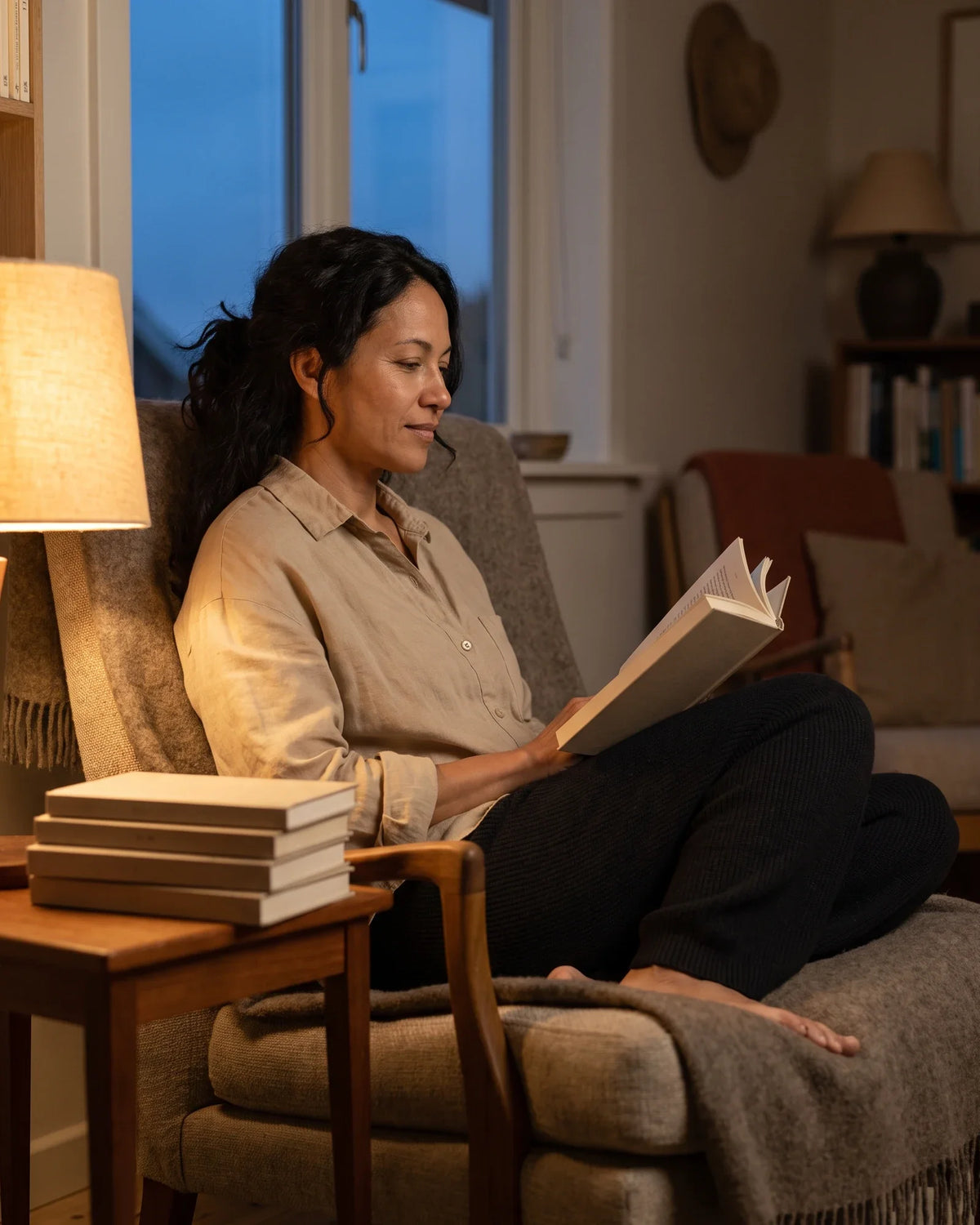 A woman reads in an armchair by lamplight with dusk in the window.
