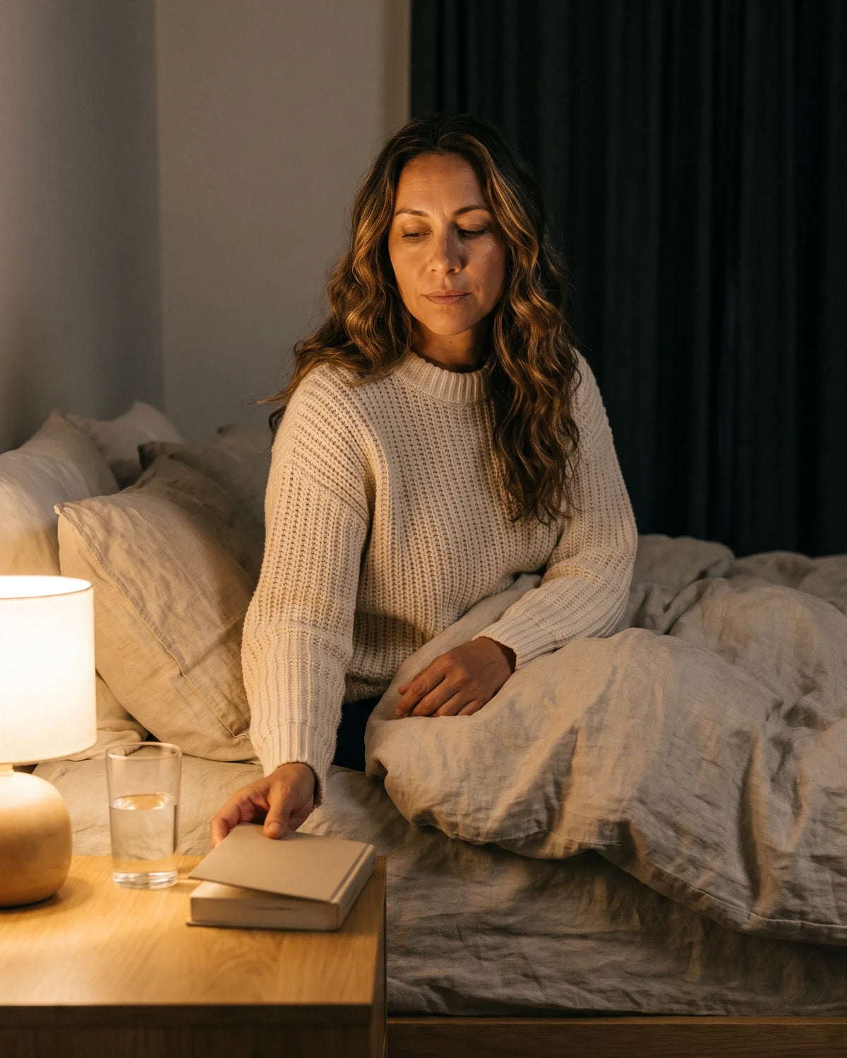 A woman sitting on her bed sets a book down on the nightstand by lamplight.
