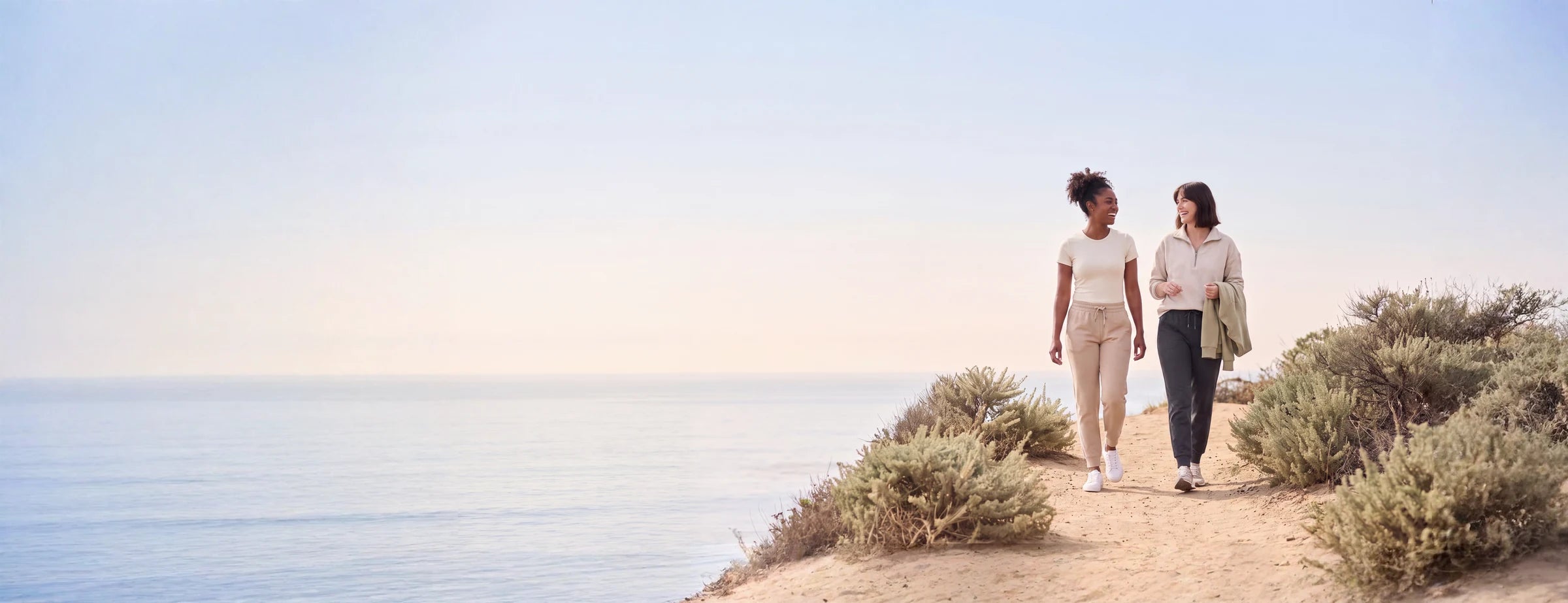 Two friends laughing as they walk a sandy coastal path above the ocean in morning light.